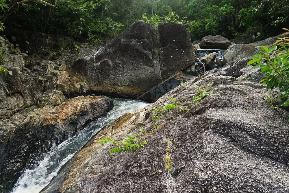 Water running over broad granite boulders at Than Sadet waterfall, surrounded by dense forest.