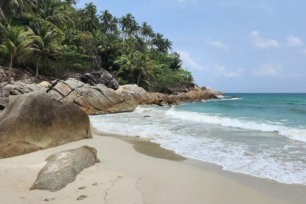 Granite boulders on the sand at Bottle Beach, with turquoise surf and a wall of palms behind.