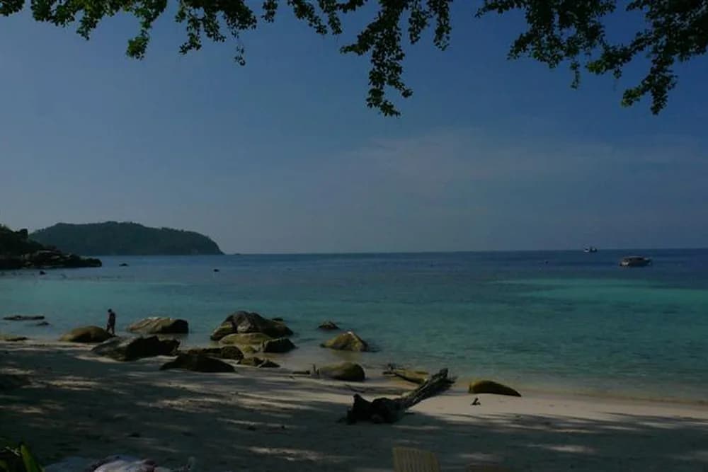 Haad Khom framed by overhanging branches, calm clear water and rocks along the shoreline.